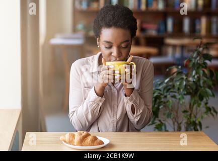 Hübsche afroamerikanische Frau, die ihren Morgenkaffee mit Croissant genießt Im städtischen Café Stockfoto