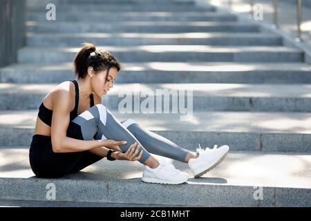 Afrikanische Frau in Sportkleidung sitzt auf der Treppe, massiert ancle Stockfoto
