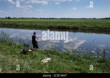 Fischer mit Zigarette im Mund steht mit einer Angelrute am Ufer eines kleinen Flusses. Wolken spiegeln sich im Wasser in typisch holländischer Landschaft Stockfoto