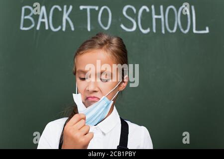 Selektiver Fokus der unzufrieden Schulmädchen berühren Schutzmaske in der Nähe Kreidetafel Mit Schultext Stockfoto