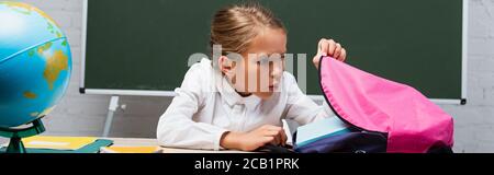 Panorama-Aufnahme von überrascht Schulmädchen Blick in Rucksack während sitzen Am Schreibtisch in der Nähe von Globe und Tafel Stockfoto