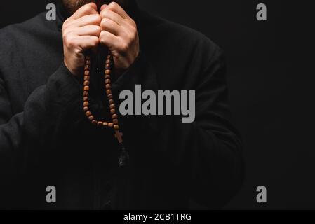 Beschnittene Ansicht des Priesters hält Rosenkranz Perlen in den Händen und Gebet isoliert auf schwarz Stockfoto