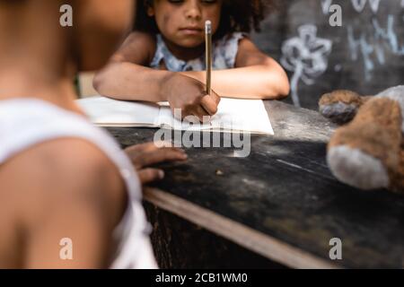 Beschnittene Ansicht des armen afroamerikanischen Kindes, das auf Papier schreibt Bruder in der Nähe Stockfoto