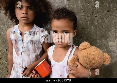 Arme afroamerikanische Kinder in schmutziger Kleidung mit Metallbecher Und Teddybär, der die Kamera in der Nähe einer Betonwand im Freien betrachtet Stockfoto