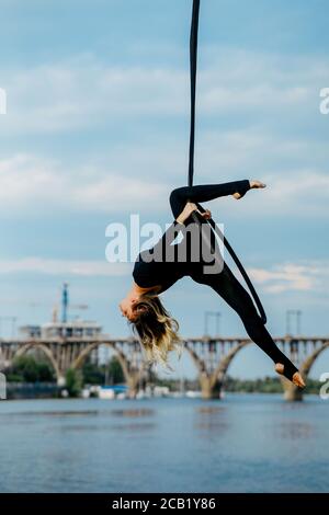 Frau Aerialistin führt akrobatische Elemente in hängenden Luftreifen vor dem Hintergrund des Flusses, Brücke und blauen Himmel. Stockfoto
