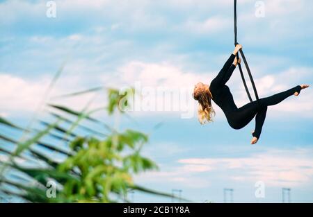 Frau Aerialistin führt akrobatische Elemente in hängenden Luftreifen vor dem Hintergrund des blauen Himmels, weiße Wolken und Blätter. Speicherplatz kopieren. Stockfoto