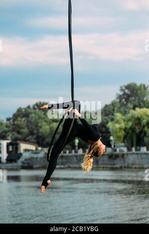 Frau Aerialistin führt akrobatische Elemente in hängenden Luftreifen vor dem Hintergrund des Flusses, Bäume und blauen Himmel mit Wolken. Stockfoto