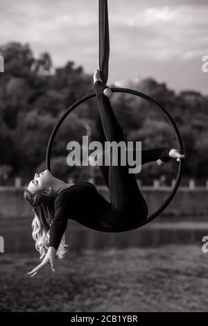 Frau Aerialistin führt akrobatische Elemente in hängenden Luftreifen vor dem Hintergrund des Flusses, Bäume und Himmel mit Wolken. Schwarzweiß-Bild. Stockfoto
