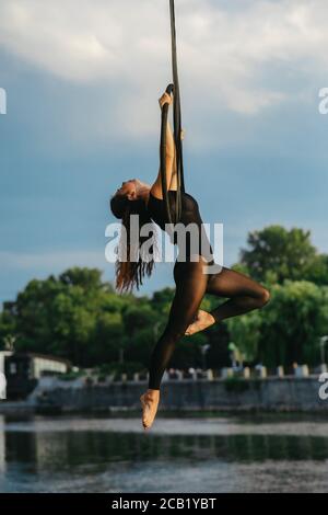 Frau Aerialistin führt akrobatische Elemente in hängenden Luftreifen vor dem Hintergrund des Flusses, Bäume und blauen Himmel mit Wolken. Stockfoto