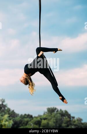 Frau Aerialistin führt akrobatische Elemente in hängenden Luftreifen vor dem Hintergrund des blauen Himmels, weiße Wolken und Bäume. Stockfoto