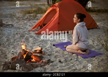 Charmante junge Frau, die am Lagerfeuer sitzt und Kaffee trinkt Stockfoto