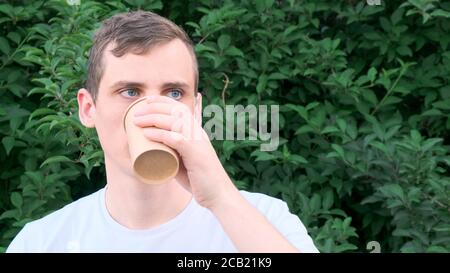 Mann mit blauen Augen trinkt Kaffee aus einem Pappbecher Auf dem Hintergrund der Büsche Stockfoto