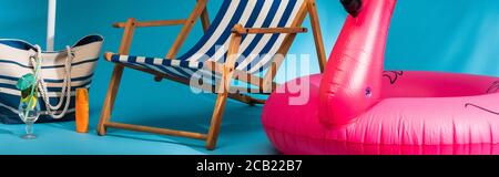 Nahaufnahme des gestreiften Liegestuhls in der Nähe von aufblasbarem Flamingo, Sonnencreme, Strandtasche und Cocktail auf blauem Hintergrund, Panoramaaufnahme Stockfoto