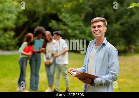 Student Guy Vorbereitung Für Prüfung Im Freien, Während Seine Freunde Chatten Im Hintergrund Stockfoto