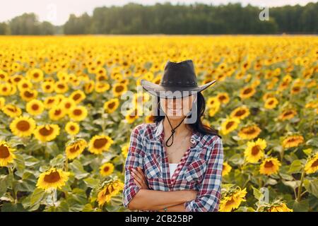 Junge Frau Agronomin trägt Cowboyhut, kariertes Hemd und Jeans auf Sonnenblumenfeld. Umfassendes Erntekonzept. Stockfoto