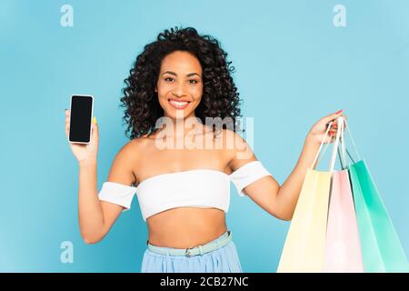 Lächelnde afroamerikanische Frau hält Einkaufstaschen und Smartphone mit Leerer Bildschirm auf blau isoliert Stockfoto