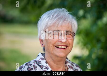 Kopfportrait einer 79-jährigen Frau mit weißen Haaren vor dem verschwommenen Hintergrund eines Stadtparks, fröhlich lachend vor der Kamera. Stockfoto