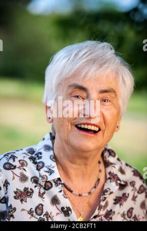 Kopfportrait einer 79-jährigen Frau mit weißen Haaren vor dem verschwommenen Hintergrund eines Stadtparks, fröhlich lachend vor der Kamera. Stockfoto