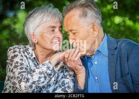 Kopf- und Schulteransicht eines älteren Rentnerpaares, das die Hand vor grünem verschwommenem Hintergrund küsst. Stockfoto