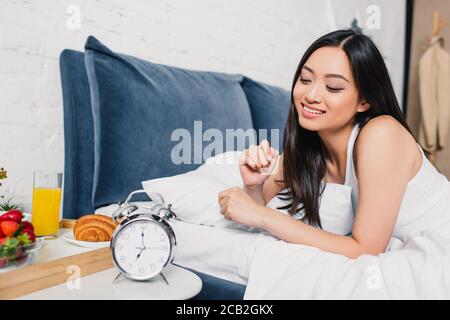 Selektive Fokus der lächelnden asiatischen Mädchen auf dem Bett in der Nähe liegen Frühstück und Wecker am Nachttisch Stockfoto