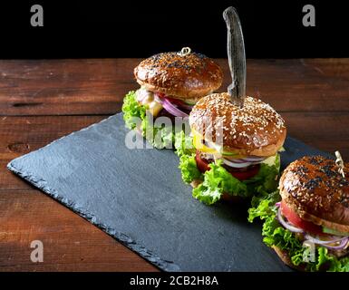 Drei leckere Burger mit gebratenem Rinderschnitzel, Tomate, Salat und Zwiebeln, knusprigem Weizenmehl-Brötchen mit Sesam. Fastfood auf einem hölzernen Wildschwein Stockfoto