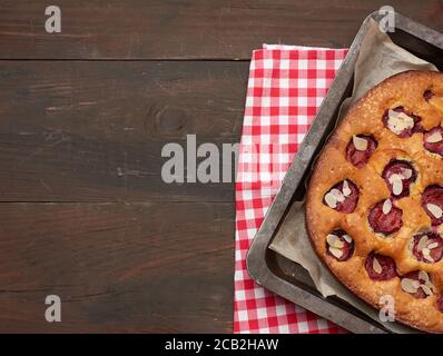 Gebackener Biskuitkuchen mit Pflaumen in einem Blech auf braunem Holzhintergrund, Draufsicht, Kopierraum Stockfoto