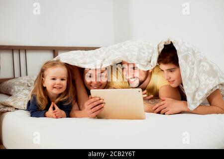Freundliche kaukasische Familie, die morgens zu Hause unter der Decke liegt, Videos auf dem Tablet ansieht, Mutter Vater und Kinder im Bett in legerer Kleidung Stockfoto