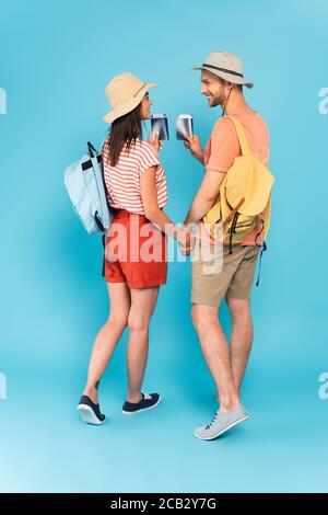 Glückliches Paar mit Hüten, die beim Suchen Hände und Pässe halten Aufeinander auf blau Stockfoto