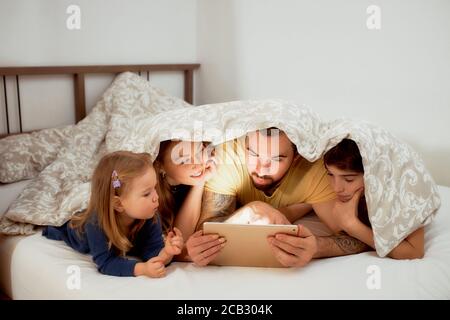 Schöne Familie bestand aus jungen Eltern und niedlichen Kindern Tochter und Sohn genießen Sie die Zeit zusammen zu Hause, auf dem Bett unter der Decke liegen Stockfoto