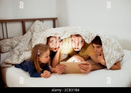 Schöne kaukasische Familie, die morgens zu Hause unter der Decke liegt, Videos auf dem Tablet ansieht, Mutter Vater und Kinder im Bett in legerer Kleidung Stockfoto