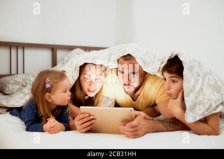 kaukasische Familie, überrascht Mutter Vater Sohn und Tochter Blick auf den Bildschirm des Tabletts, interessante Film oder Video, liegend unter Bettdecke auf dem Bett Stockfoto