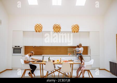 Tagsüber mit der Familie zu Hause. Vater arbeitet von zu Hause aus mit einem Laptop. Mutter kocht, während sie das Baby hält. Stockfoto