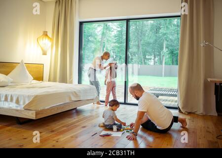 Panoramafenster im Zimmer. Junge Eltern mit ihren Kindern im Zimmer. Mutter und Tochter, Vater mit Sohn, der auf dem Boden spielt. Wachsende Generation, Familie Stockfoto