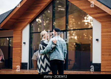 Kaukasische Frau und Mann stehen umarmt neben einem großen Haus auf dem Land. Geschieht durch den Kauf eines Hauses Stockfoto