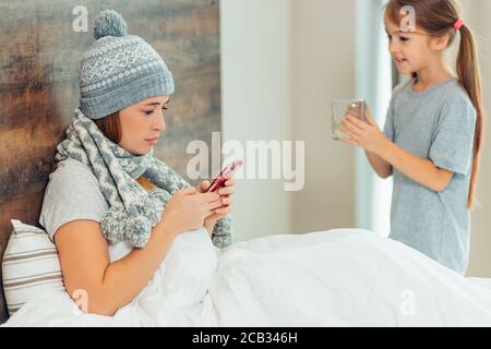 Nette kaukasische Kind Mädchen bringt heißen Tee zu kranken Mutter liegend auf dem Bett in Hut und Schal, vorsichtig Kind Mädchen und kranke Mutter Stockfoto