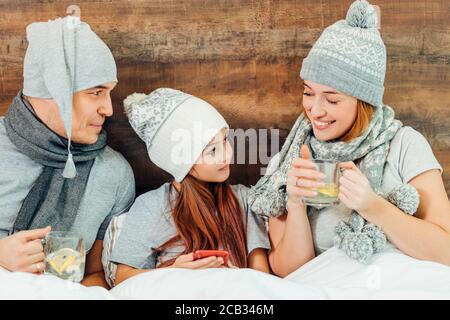 Porträt der schönen kaukasischen Familie leiden an Krankheit, trinken heißen Tee Behandlung von Krankheiten, tragen Hüte, Tücher und Hauskleidung, sitzen Stockfoto
