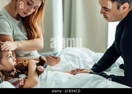 Familienkonzept. kaukasische Frau überprüfen Temperatur der süßen Tochter leiden an Kälte zu Hause, liegen auf dem Bett mit sorgfältiger Mutter und Vater Stockfoto