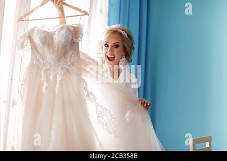 Braut Morgenvorbereitung. Glückliche schöne junge Frau in Seide Bademantel berühren ihr Hochzeitskleid, während in der Nähe des Fensters stehen Stockfoto