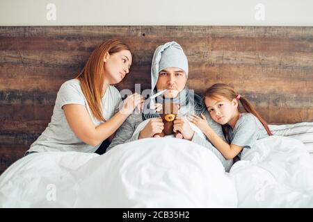 Sorgfältige Frau und Kind Mädchen sitzen auf dem Bett mit kranken Vater mit hoher Temperatur, Fieber. Frau und Kind unterstützen ihn und geben Medikamente Stockfoto