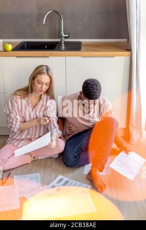 Junge Mixed-Rennen Paar von verschiedenen Studenten in Freizeitkleidung, sitzen zusammen auf Küchenboden zu Hause arbeiten bei ihrer Kursarbeit konzentriert Stockfoto