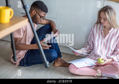 Junge Mixed-Rennen Paar von verschiedenen Studenten in Freizeitkleidung, sitzen zusammen auf Küchenboden zu Hause arbeiten bei ihrer Kursarbeit konzentriert Stockfoto