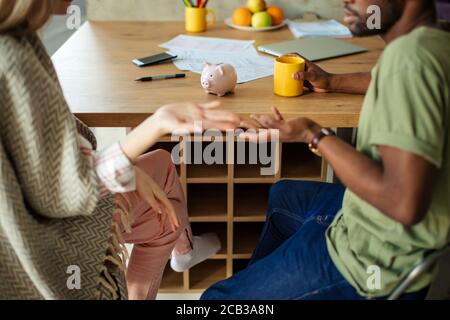 Die abgeschnittene Aufnahme des multiethnischen Paares hat einige Missverständnisse, drücken Sie diesen Zustand durch verdutzte Handbewegung aus, während Sie in Livi am Tisch sitzen Stockfoto