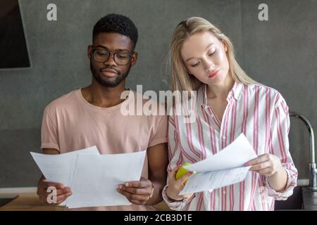 Junge gemischte Rennen Paar von verschiedenen Studenten in Freizeitkleidung, sitzen Seite an Seite, beschäftigt mit dem Lesen der Kursarbeit. Clevere multirassische Freunde studieren Stockfoto