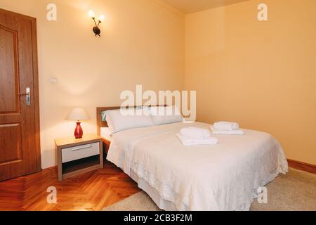 Großes Queen-Size-Bett mit Kissen und flauschigen Handtüchern auf der Tagesdecke in einem geräumigen Zimmer mit Lese- und Wandlampen und Fußmatte. Stockfoto