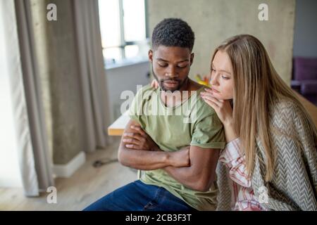 Kaukasische blonde Frau Konsolen und unterstützt ihre afrikanische Hipster Freund, der mit düsteren und frustrierten Blick, Paar sitzt in gemütlichen Leben Stockfoto