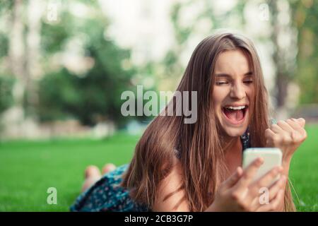 Aufgeregt schöne Mädchen erhalten eine sms-Nachricht mit guten Nachrichten in einem Handy außerhalb im Park liegend auf grünen Rasen Bäume auf dem Hintergrund. Mehrzul Stockfoto