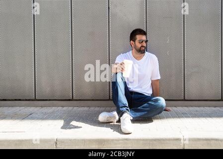 Bärtiger Kerl mit Sonnenbrille auf dem Boden sitzend, während er ein hält Kaffee mitnehmen Stockfoto