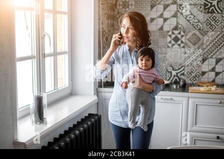 Beschäftigt ernsthafte Mutter hält ihr Baby und sprechen auf dem Smartphone in der Küche. Nahaufnahme Foto, kostenlos, Freizeit Stockfoto