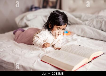 Schönes Baby Mädchen liegt auf dem Bett und liest ein Buch zu Hause, Nahaufnahme Foto, neue Generation, Baby bekommt Wissen Stockfoto