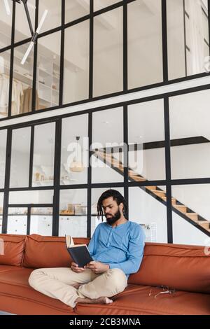 Schöner lächelnder junger afrikanischer Mann, der zu Hause auf einer Ledercouch sitzt und ein Buch liest Stockfoto